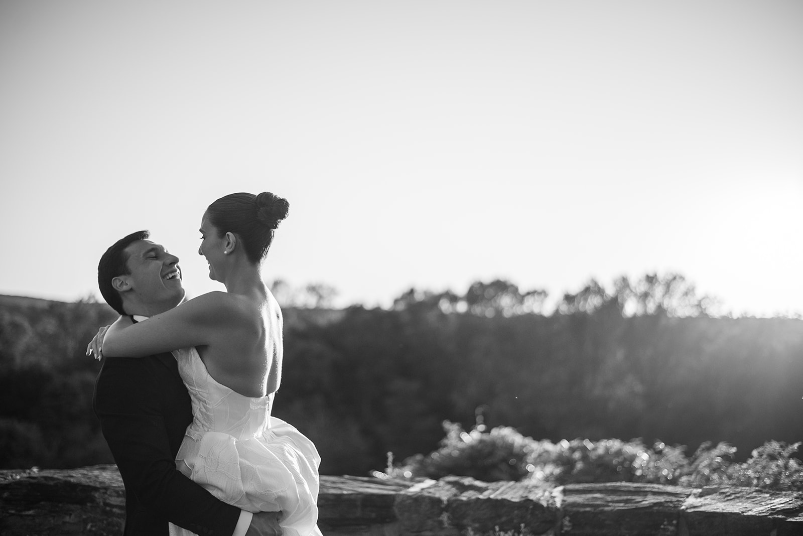 Black and white film photo of the groom lifting and laughing with his bride by a stone wall at sunset.